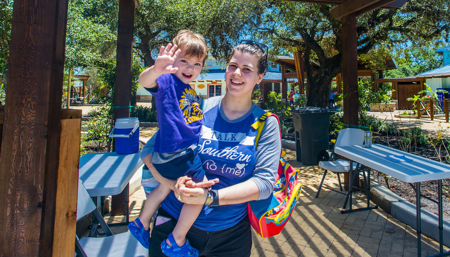 BLOG A woman holding a young child who is waving, both standing outdoors on a sunny day with tables and chairs around.