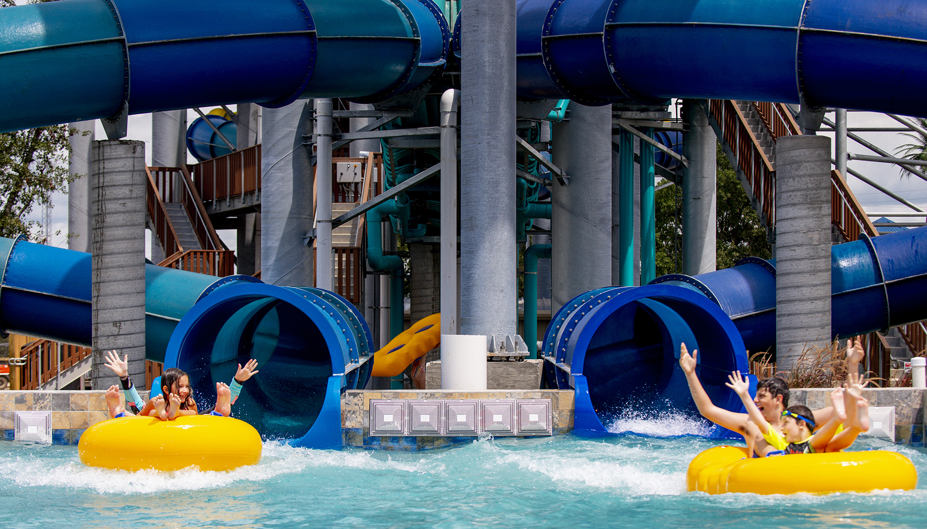 BLOG People riding yellow inner tubes emerge joyfully from water slides into a pool at an outdoor water park.