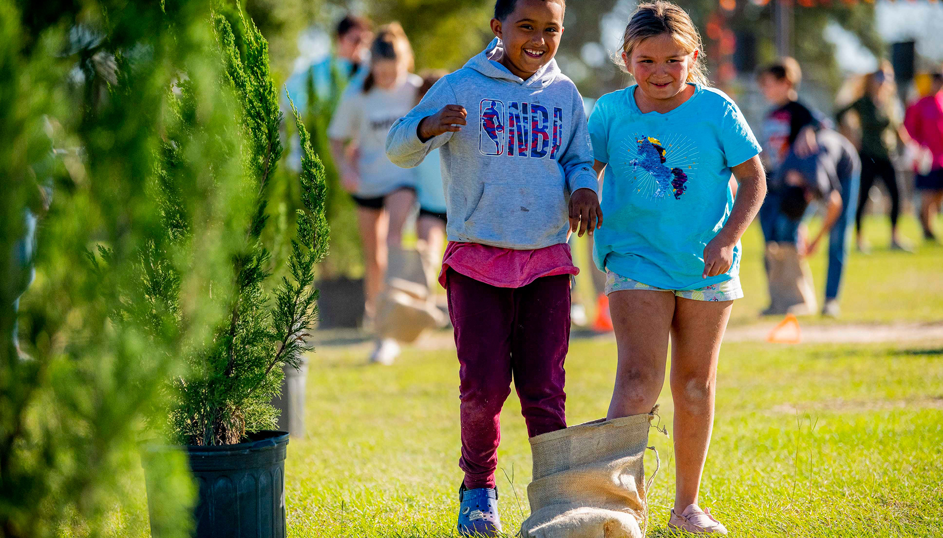 BLOG Two children participate in a sack race during an outdoor event, both smiling and holding hands, surrounded by greenery and other participants in the background.