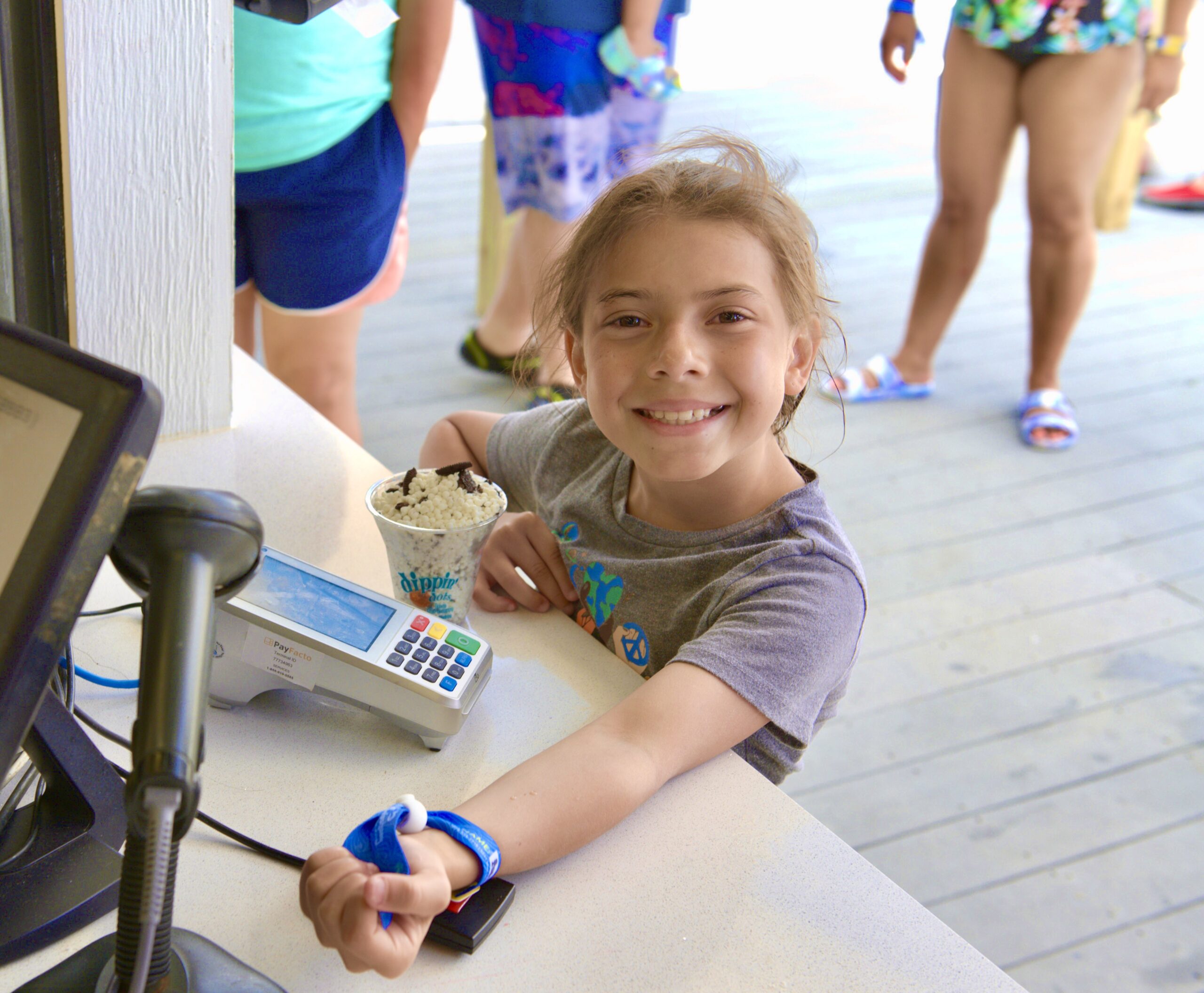 splash cash Young girl smiling and holding her wristband up while standing at a counter with a card reader. She has a dessert bowl filled with ice cream and toppings next to her.