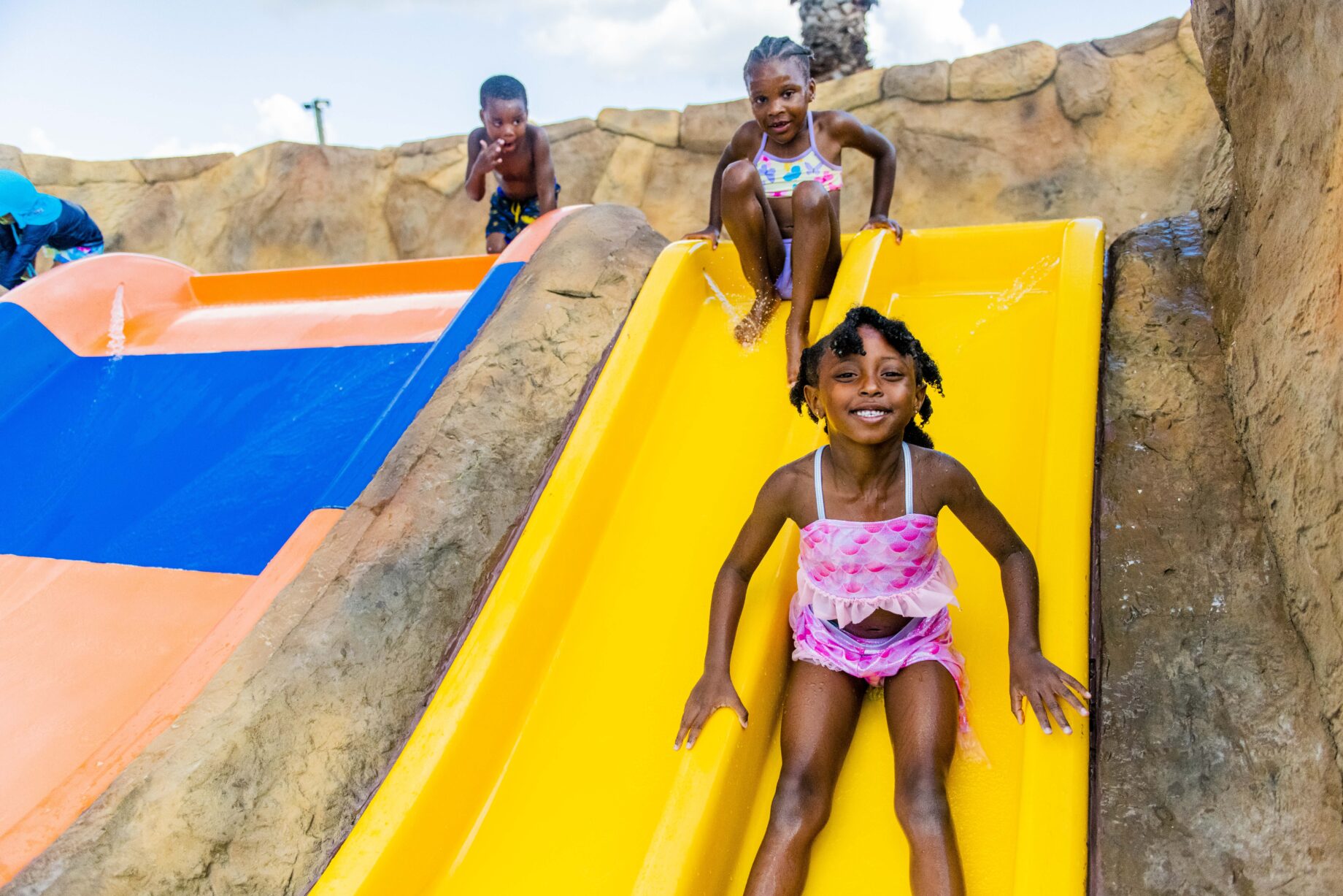 Splashway_2007 Children in swimsuits slide down yellow and orange slides at a water park. They are smiling and enjoying themselves on a sunny day.