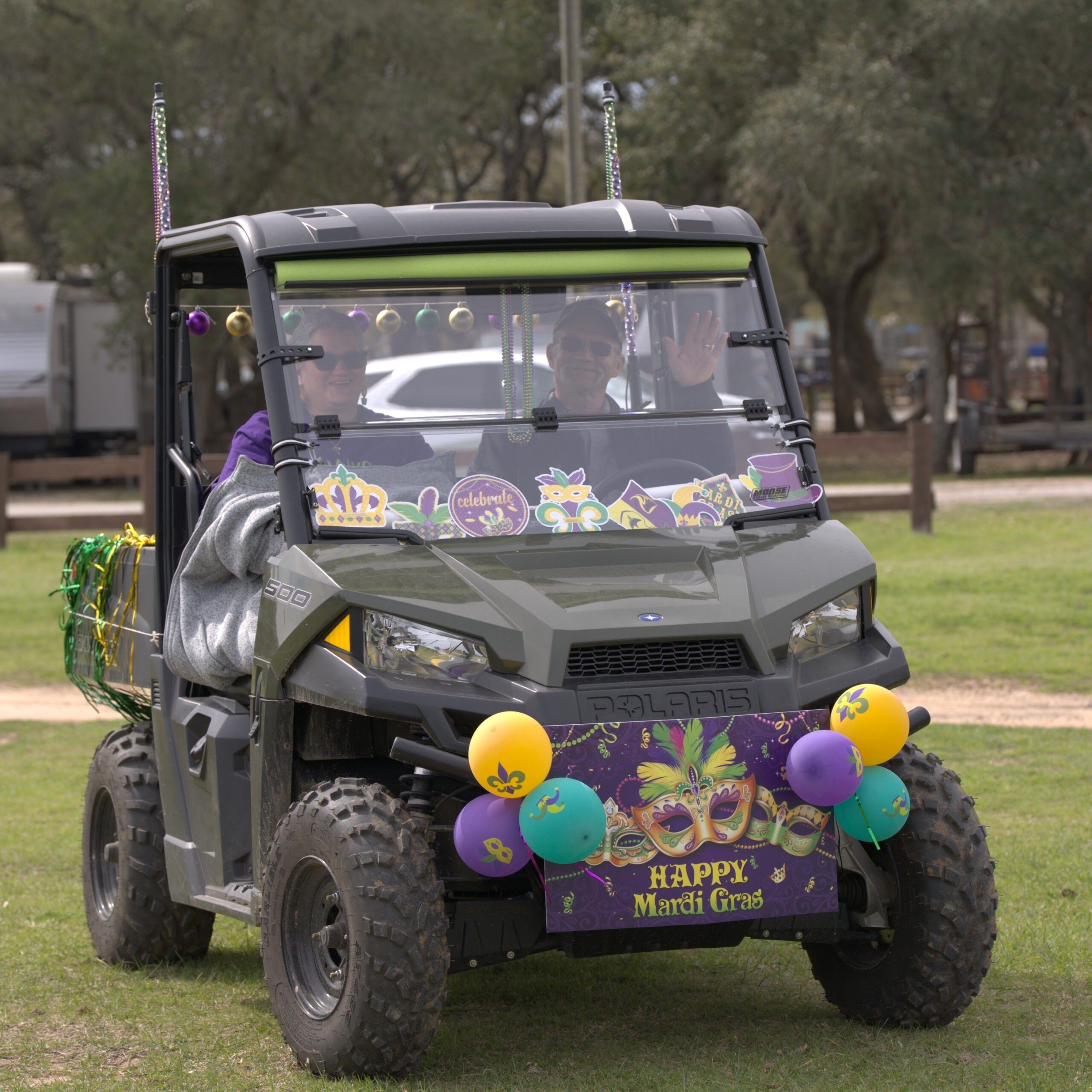 NS3Gs9X2 A person drives a decorated off-road vehicle with Mardi Gras banners and balloons on a grass field, waving and wearing sunglasses.
